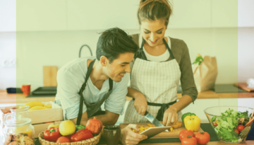 Ernährungsberatung: Zwei junge Menschen bereiten fröhlich gesunde Mahlzeiten zu, während sie eine Tablet-Anwendung verwenden, um nach Rezepten zu kochen und ihre Kalorien im Blick zu behalten.