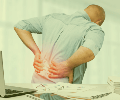 Ein Mann dreht sich vom Schreibtisch weg und stützt sich mit den Händen auf den schmerzenden Rückenbereich. Ein roter Punkt symbolisiert Rückenschmerzen.